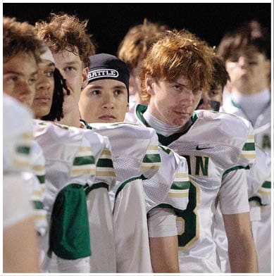 A group of people wearing white and green sports uniforms stand closely together, with one person in the center wearing a black headband labeled “BATTLE." Description generated by AI