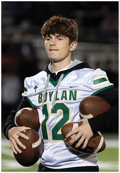 A person wearing a white and green football jersey with the number 12 is holding three footballs while standing on a field. Description generated by AI