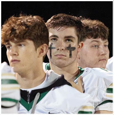 Two people wearing sports jerseys are standing close together, with one person having black crosses painted on their cheeks. Description generated by AI