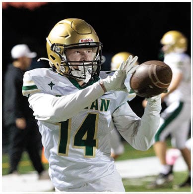 A person wearing a gold football helmet and white uniform with the number 14 is catching a football on a field with other players and a coach in the background. Description generated by AI