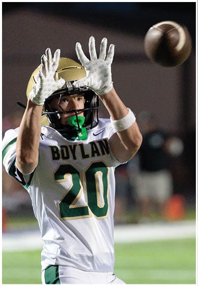 A person wearing a football uniform with the number 20 is reaching up with both hands to catch a football in midair on a field. Description generated by AI