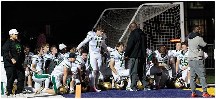 A group of people in sports uniforms are gathered on the sidelines of a football field near the end zone, some sitting and some standing, with helmets on the ground. Description generated by AI