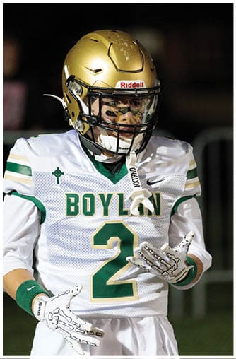 A person wearing a gold football helmet with a clear visor and a black faceguard is looking intently to the side against a dark background. Description generated by AI