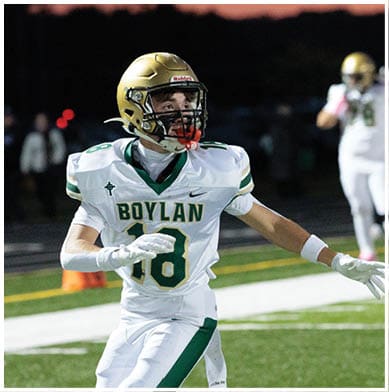 A person wearing a white and green football uniform with a gold helmet is running on a field during sunset. Description generated by AI