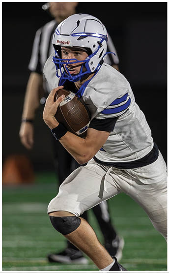 A person wearing a white and blue football uniform is running while holding a football tightly with both hands on a field, with a referee standing in the background. Description generated by AI
