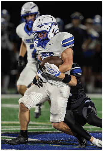 Two people in white and blue football uniforms and helmets are engaged in a game, with one holding a football tightly. Description generated by AI