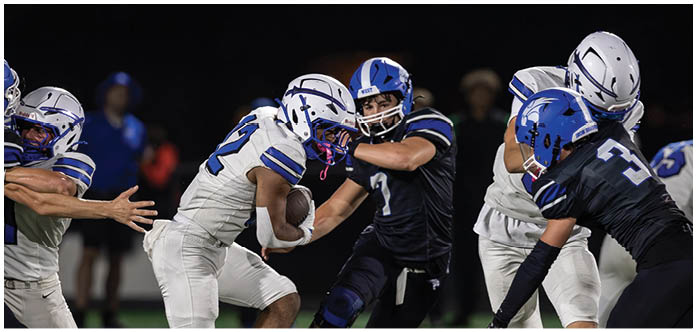 Players in white and blue football uniforms are engaged in a play on a nighttime field with one player holding the ball while others attempt to block or tackle. Description generated by AI