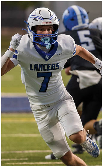 A person wearing a white football uniform with the number 7 and the word Lancers is running on a football field while other players in white and black uniforms are engaged in a play behind them. Description generated by AI