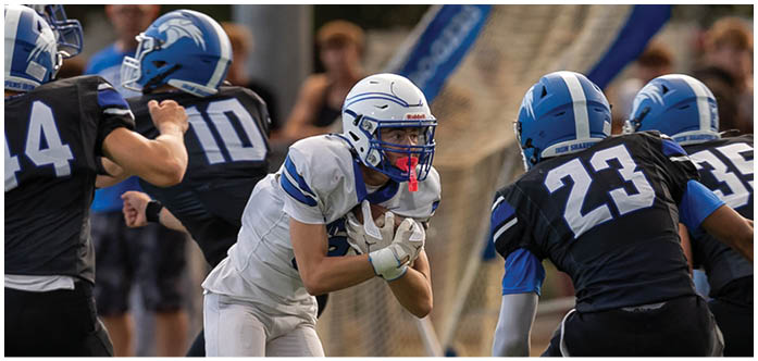 A group of people wearing football uniforms are actively engaged in a play on a grassy field with spectators in the background. Description generated by AI
