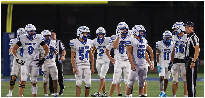 A group of football players in white uniforms and helmets stand on a field near a referee dressed in black and white stripes. Description generated by AI