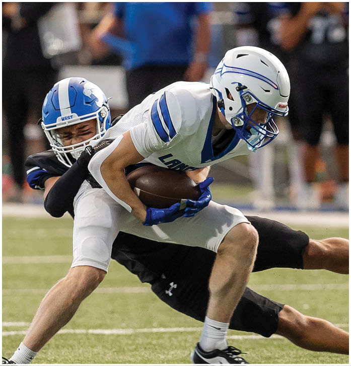 Two people in football uniforms are engaged in a tackle on a grassy field with a crowd and team members in the background. Description generated by AI