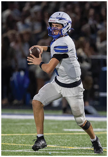A person wearing a white and blue football uniform and helmet is holding a football in a throwing position on a field with a blurred crowd in the background. Description generated by AI