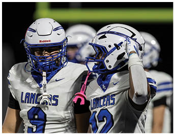 Two people wearing white football uniforms with blue accents and helmets stand close together on a field with a goalpost in the background. Description generated by AI