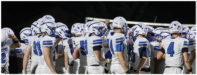 A group of people wearing white and blue football uniforms and helmets are gathered closely together outdoors at night. Description generated by AI