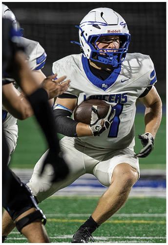 A person in white football gear is running with the ball while being approached by opponents in dark uniforms on a football field. Description generated by AI