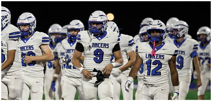 A group of people wearing white and blue football uniforms with helmets are standing together on a field at night. Description generated by AI