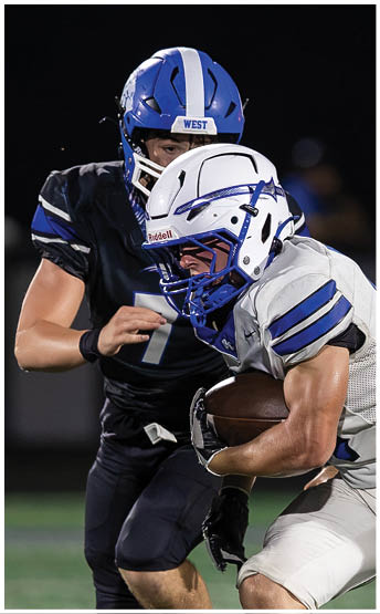 Two people in football uniforms are engaged in a play on a field at night, with one person in white running while holding the ball and the other in black attempting to tackle. Description generated by AI