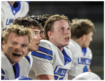 A group of people in white and blue sports uniforms sit on a bench, with one person gesturing with their hand while others look on attentively. Description generated by AI