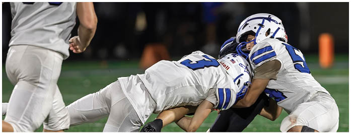 Two people in white football uniforms tackle a person in a dark uniform on a green field while another person in white runs nearby. Description generated by AI