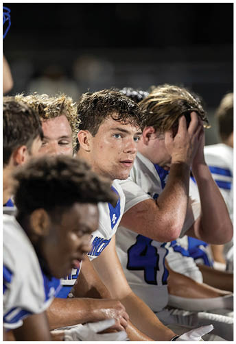 People in football uniforms sit closely together on a bench, appearing focused and contemplative during a game or practice. Description generated by AI