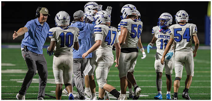 Several people in football uniforms and helmets are standing together on a field, with some wearing white jerseys with blue numbers and one person in a blue jacket. Description generated by AI