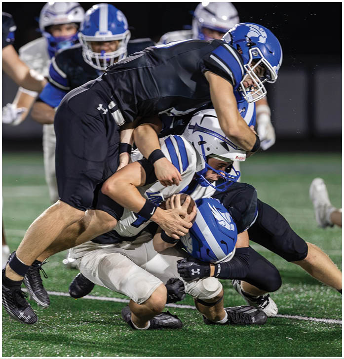 Two people in football uniforms are tackling a third person holding a football on a field during a game. Description generated by AI