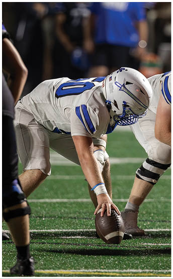 Two football players in white uniforms and helmets are positioned on the field facing an opposing player in a black uniform, preparing for a play. Description generated by AI