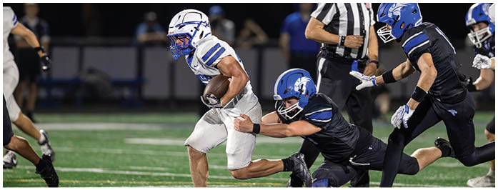 Two people in sports gear are engaged in a physical struggle on a football field near a large blue and white logo. Description generated by AI