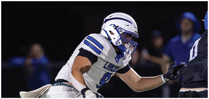 A person in a white and blue football uniform is running and reaching out toward another person in a dark uniform during a nighttime game. Description generated by AI