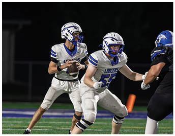 Two people wearing white and blue football uniforms and helmets are on a field, with one holding a football and preparing to throw it. Description generated by AI