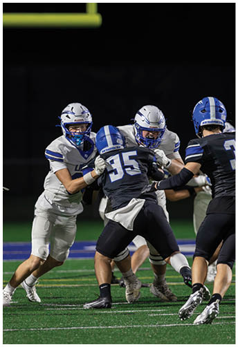 Three people wearing football helmets and uniforms are engaged in a play on a field under stadium lights. Description generated by AI