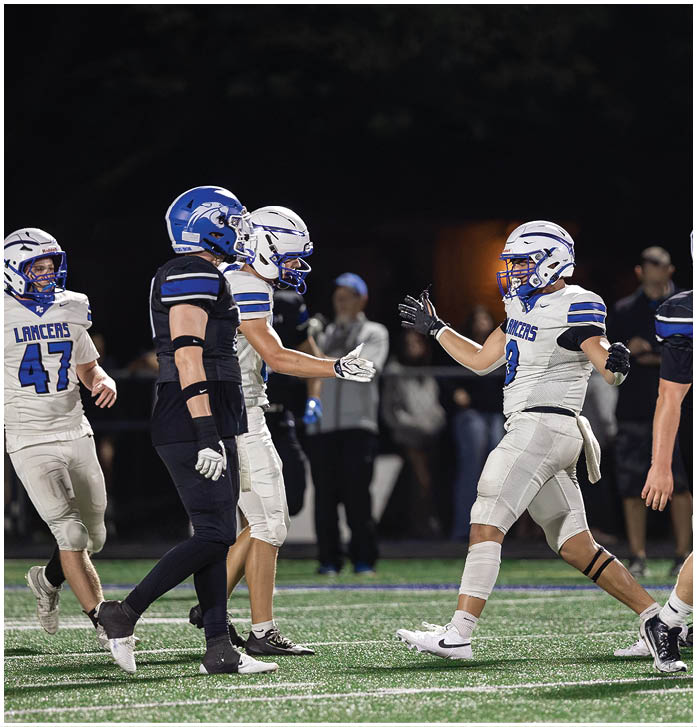 Two people wearing football helmets and uniforms are on a field at night, with one person extending a hand toward another. Description generated by AI