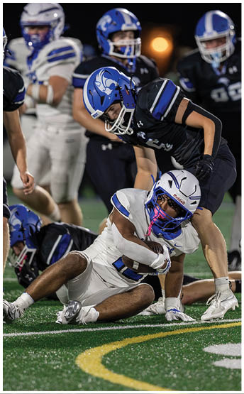 A person in white football gear is holding a football while sitting on the ground as another person in dark blue football gear leans over them during a game. Description generated by AI