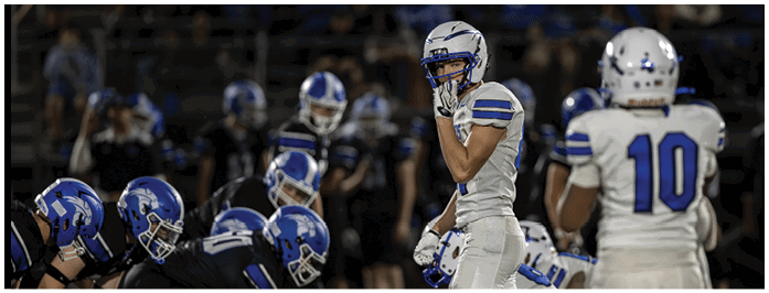 Players from two opposing football teams are lined up on the field preparing for a play, with one team in blue uniforms and the other in white uniforms. Description generated by AI