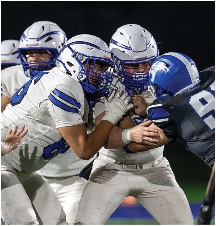 People wearing white and blue football helmets and uniforms are closely gathered, appearing to be in the middle of a game or practice. Description generated by AI