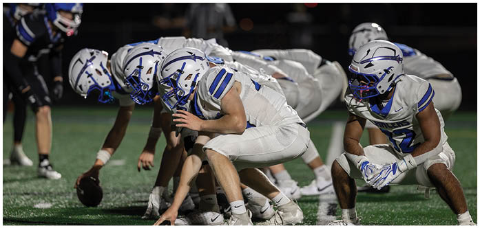 People in football uniforms and helmets are crouched in a ready position on a field, preparing for a play. Description generated by AI