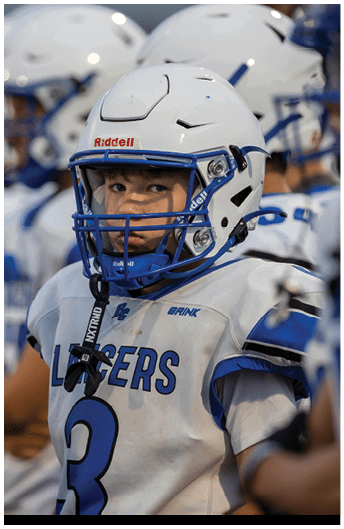 A person wearing a white and blue football uniform and helmet with a focused expression stands among others in similar gear. Description generated by AI