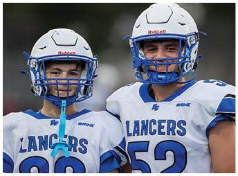 Two people wearing white football helmets with blue faceguards are smiling, with one person more prominently visible on the right side of the image. Description generated by AI