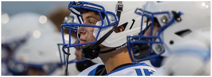 Two people wearing white helmets with blue faceguards are closely positioned, likely preparing for or participating in a football game. Description generated by AI