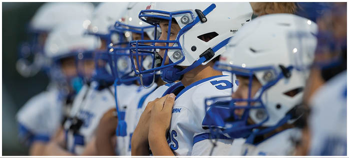 A person wearing a white football helmet with a blue faceguard is shown in close-up, with other similarly helmeted people visible in the background. Description generated by AI