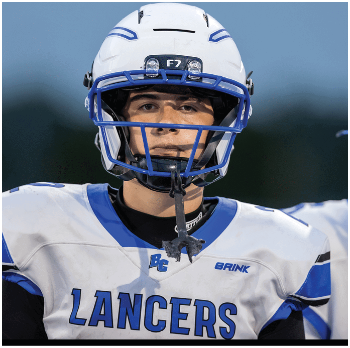 A person wearing a white and blue football uniform with the word LANCERS on the chest and a helmet with a faceguard is standing outdoors. Description generated by AI