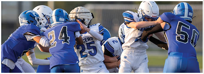 Several people wearing helmets and sports uniforms are engaged in a close physical play on a grassy field, likely during a football game. Description generated by AI