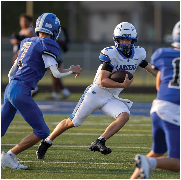 Two people in football uniforms are engaged in a play on a field, with one holding the ball and the other reaching out toward them. Description generated by AI