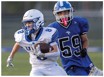 People wearing blue football uniforms are engaged in a play on a grassy field with one person extending a leg toward another player in white. Description generated by AI
