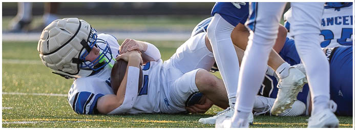 People in football uniforms are gathered on a field with one person lying on the ground and others standing or crouching nearby. Description generated by AI