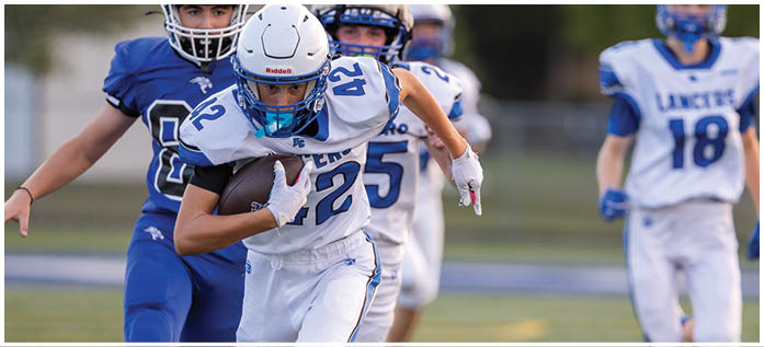 A person wearing a white and blue football uniform is running with a football while being pursued by another person in a blue uniform on a sports field. Description generated by AI