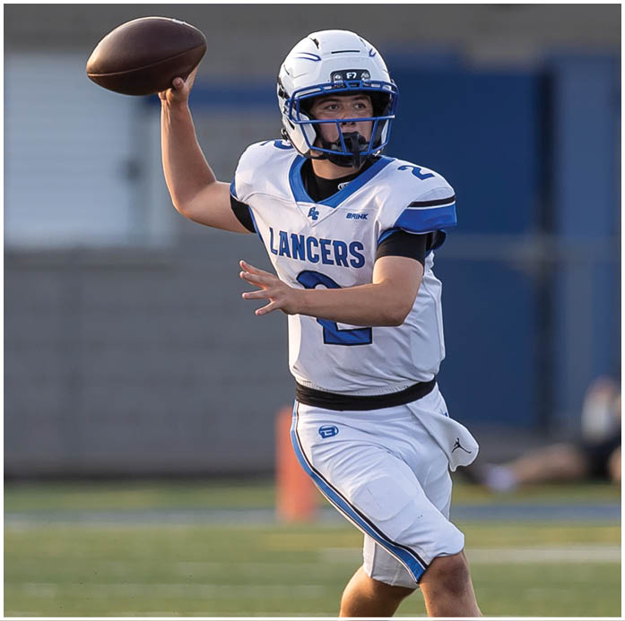 A person wearing a blue football helmet and blue jersey is standing on a field with a blurred background of a building and other people. Description generated by AI