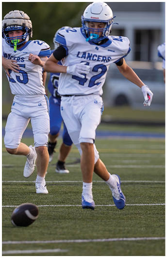 Two people in white football uniforms with blue accents are running on a field during a game. Description generated by AI