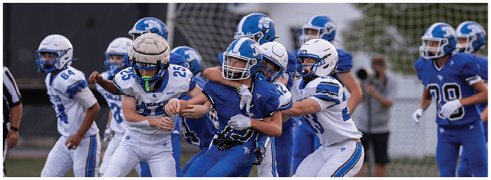 People in football uniforms are engaged in a physical play on a field with one player in blue being tackled by two players in white. Description generated by AI