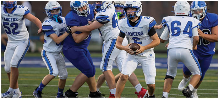 People wearing football helmets and uniforms are engaged in a game, with one person holding the ball and others blocking or tackling. Description generated by AI